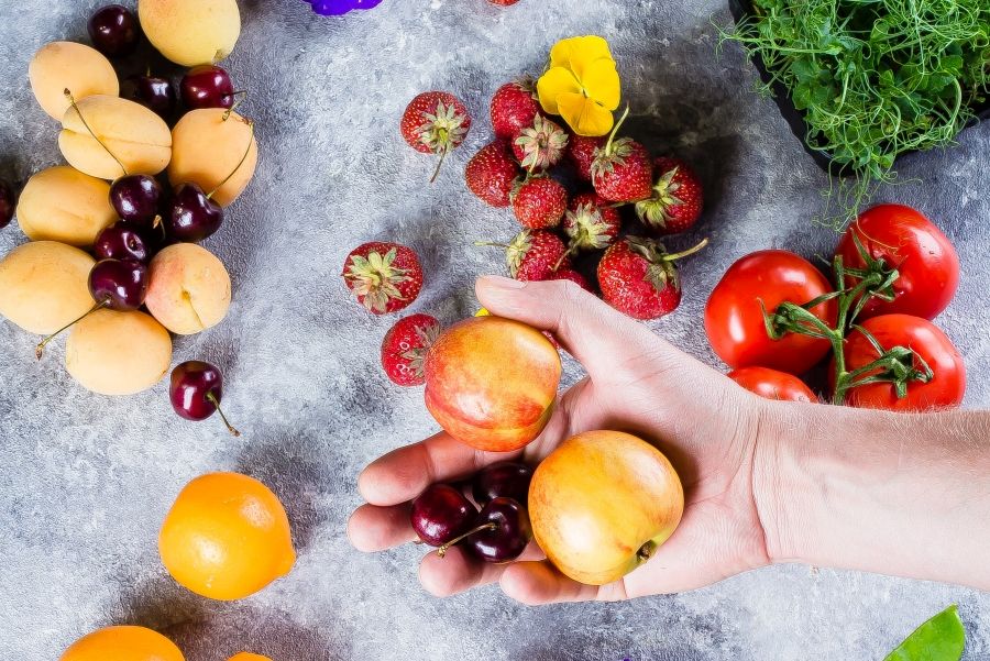 Auswahl an Obstsorten und Tomaten auf einem Tisch, eine Hand hat Pfirsische und Kirschen genommen